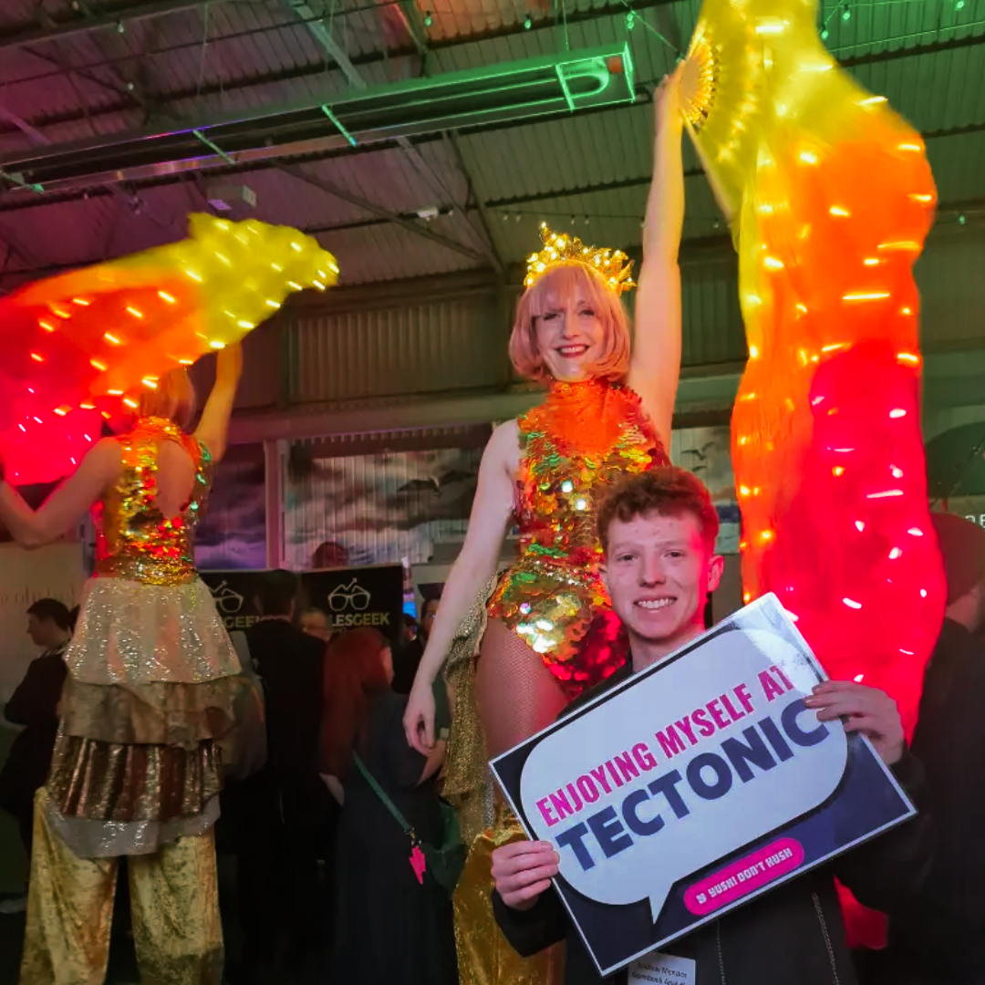Performer in LED costume next to a young man holding an Enjoying Myself At Tectonic sign