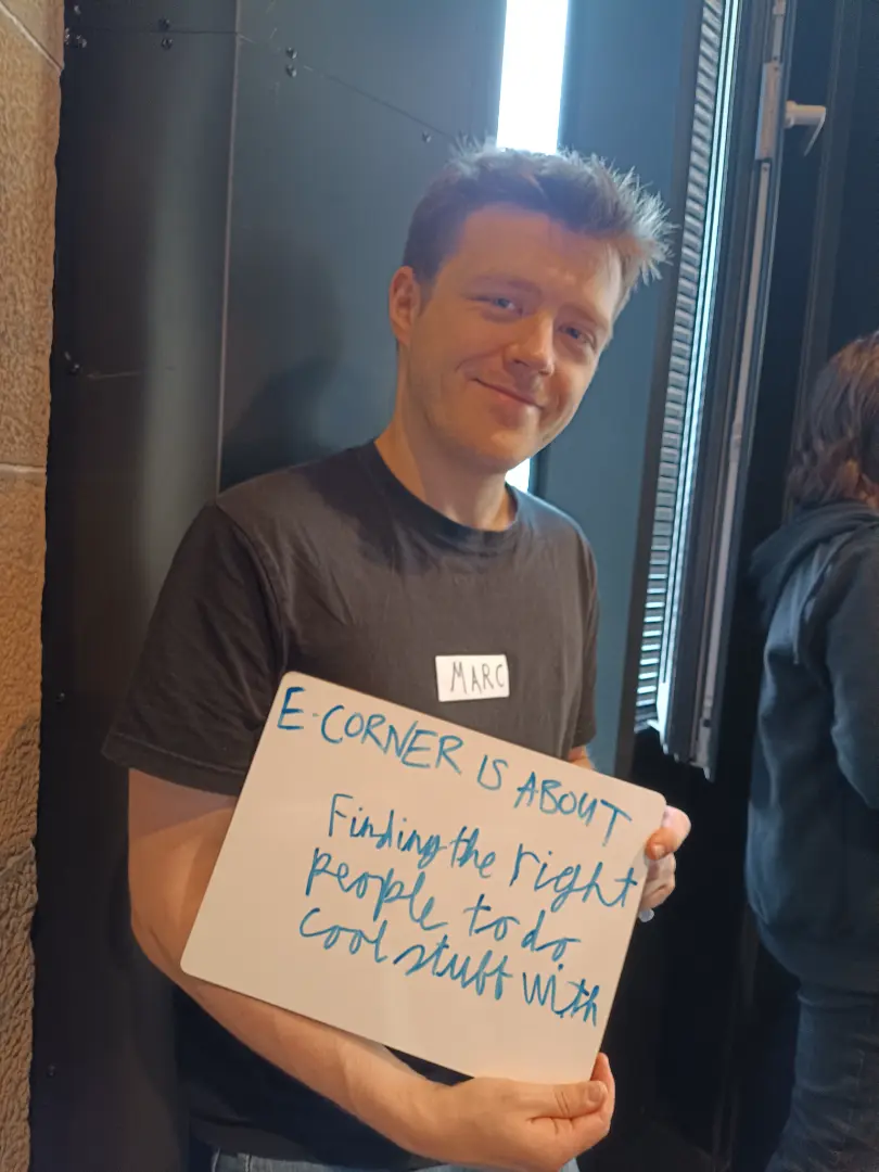A person is holding a whiteboard with handwritten text. The board reads: "E-CORNER IS ABOUT Finding the right people to do cool stuff with." The individual is smiling and has a name tag labeled "Marc."