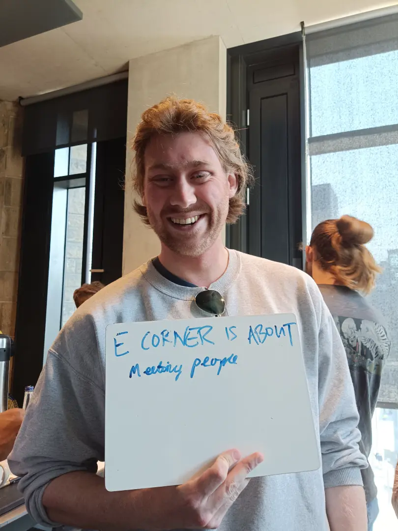 A person holding a whiteboard with the handwritten text 'E CORNER IS ABOUT Meeting people', smiling in a casual indoor setting.