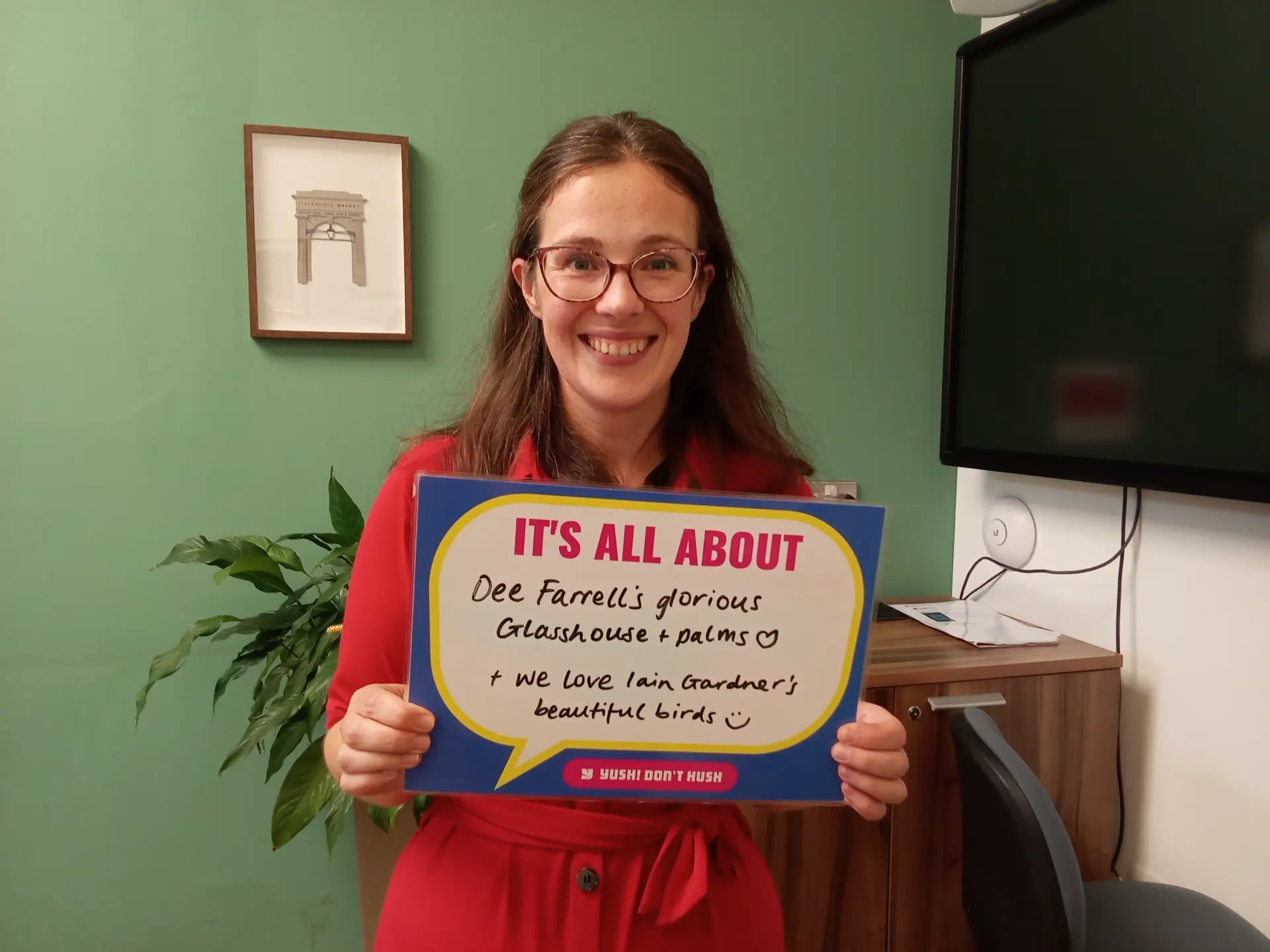 A woman is smiling while holding a sign that says 'IT'S ALL ABOUT Dee Farrell's glorious Glasshouse + palms ♡ + we love Iain Gardner's beautiful birds :)'. She is standing in an indoor setting with a green wall and a framed picture behind her.