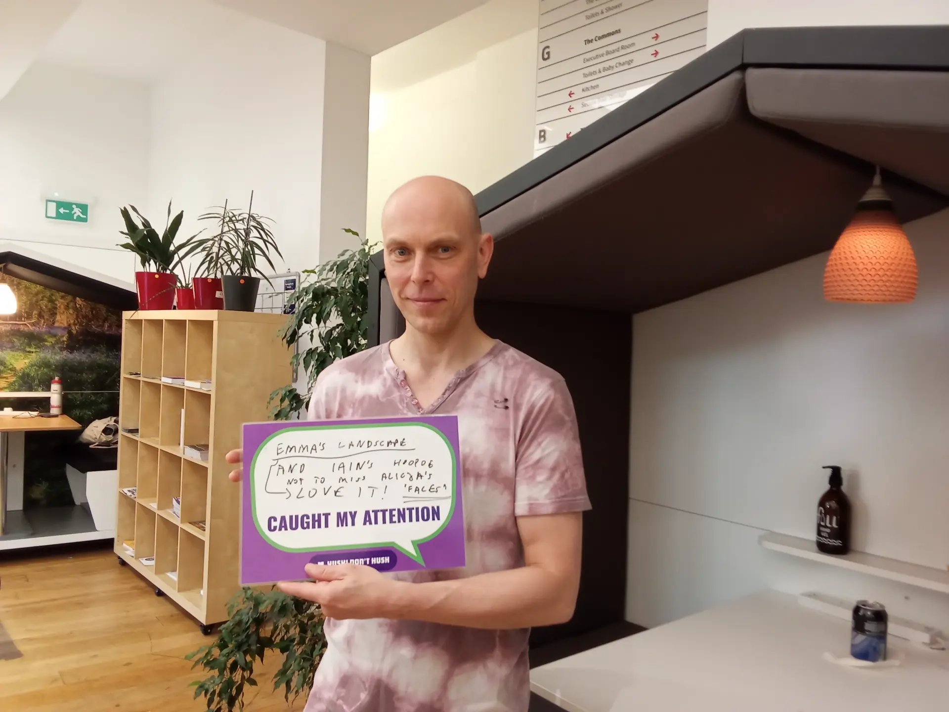A person is holding a sign with text in a cozy office setting, featuring plants and shelving in the background. The handwritten text on the sign reads: "EMMA'S LANDSCAPE AND IAIN'S HOTDOG NOT TO MISS ALISON'S CLUEY IT! 'FARCE' CAUGHT MY ATTENTION."