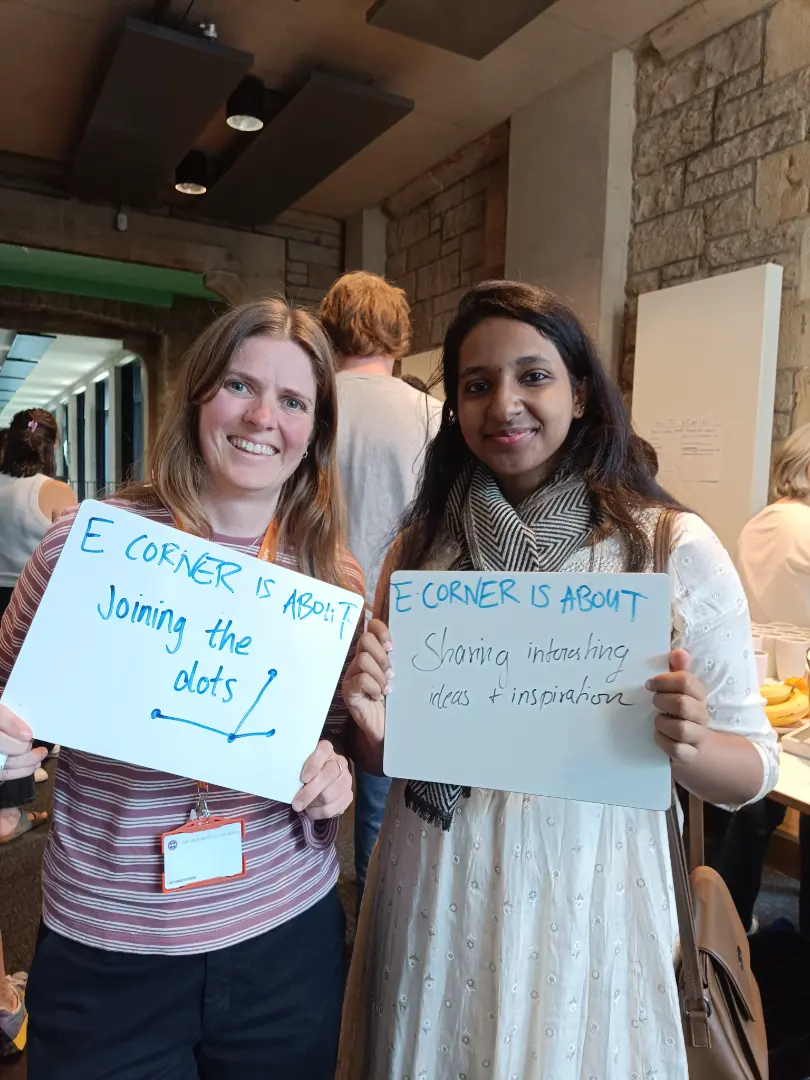 Two people are holding signs that say 'E CORNER IS ABOUT Joining the dots' and 'E CORNER IS ABOUT Sharing interesting ideas & inspiration'. They are smiling and standing in an indoor setting.