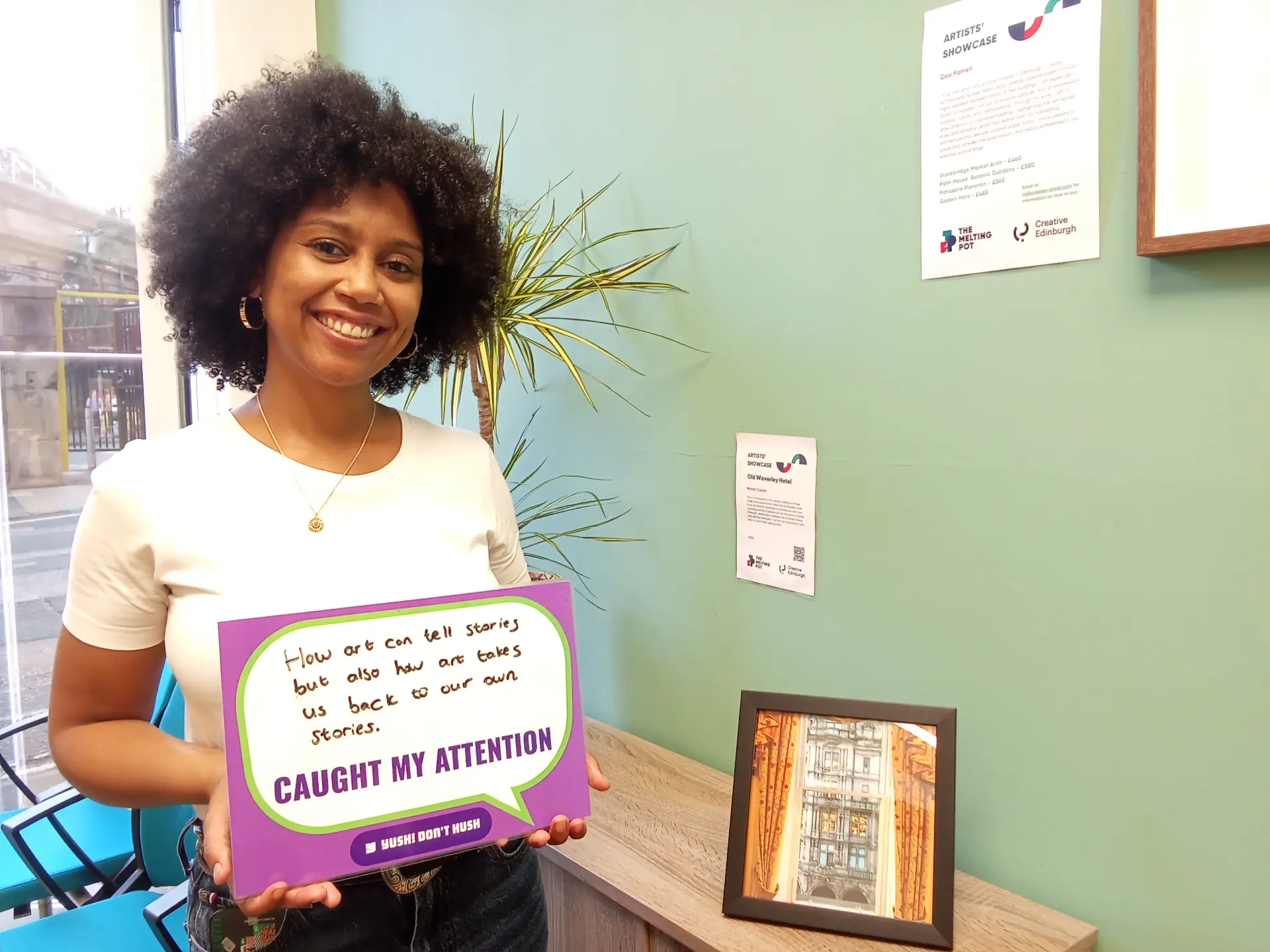 A woman is smiling while holding a sign with handwritten text. The text reads: "How art can tell stories but also how art takes us back to our own stories. CAUGHT MY ATTENTION. # YOU SHI DON'T HUSH." There are informational posters on the wall and a framed picture on a table.