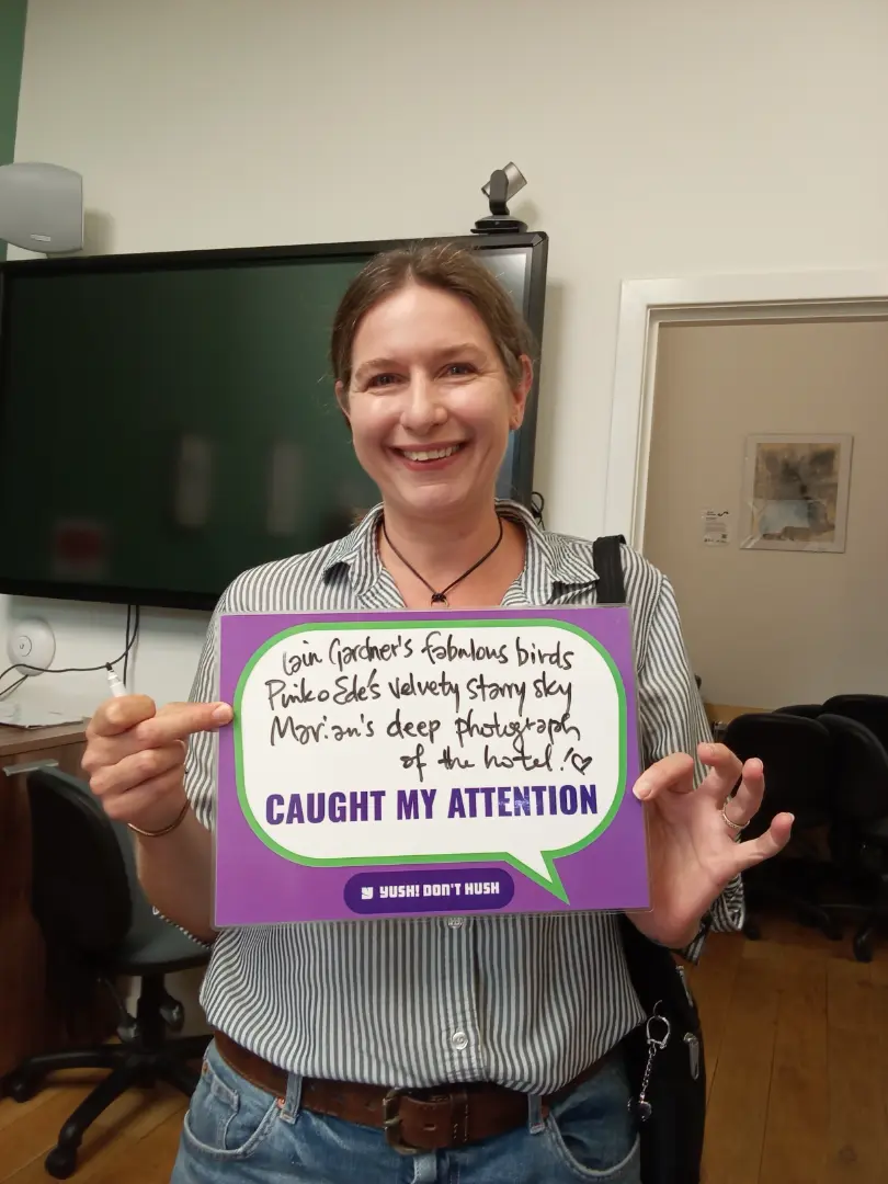 A person is smiling and holding up a sign that reads: "Gavin Gardner's fabulous birds Pinkie Edel's Velvety starry sky Miriam's deep photograph of the hostel. ♥ Caught my attention. # Hush! Don't Hush". The sentiment of the photo is positive and cheerful.