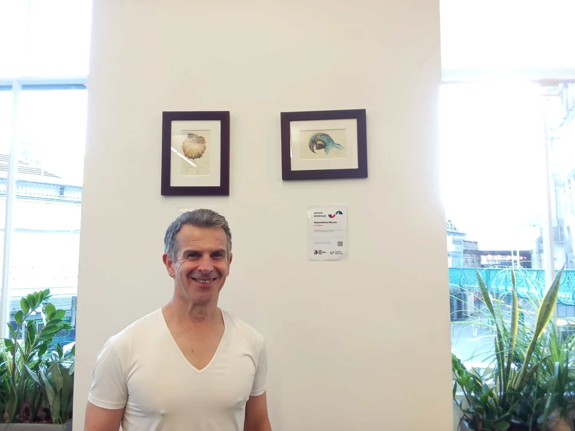 A person is standing indoors next to a wall with framed pictures and a sign. The individual is smiling and wearing a white shirt, with various plants in the background.